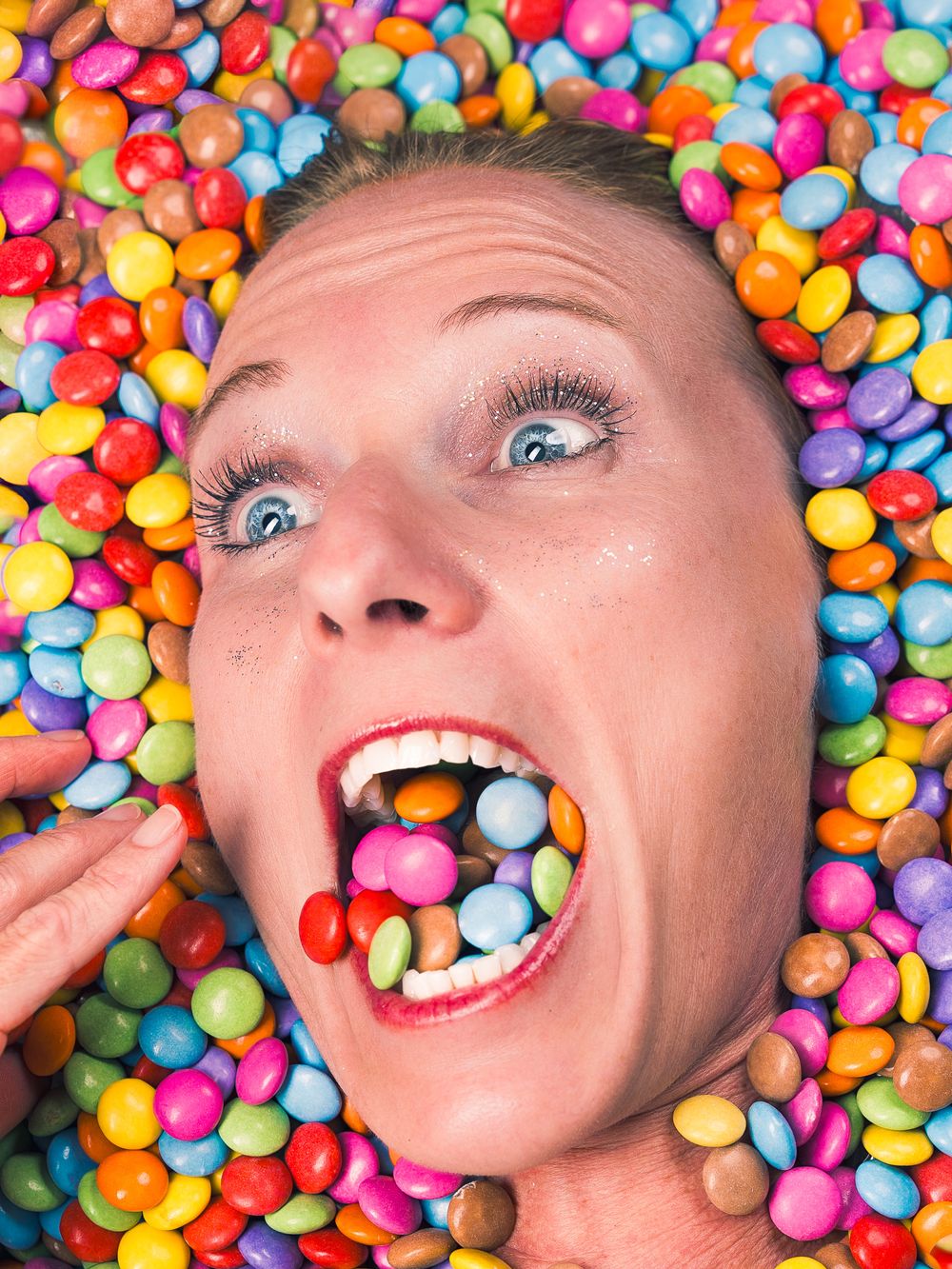 Woman lying in colorful candy-coated chocolates with mouth full of sweets, blue eyes wide open in playful expression.
