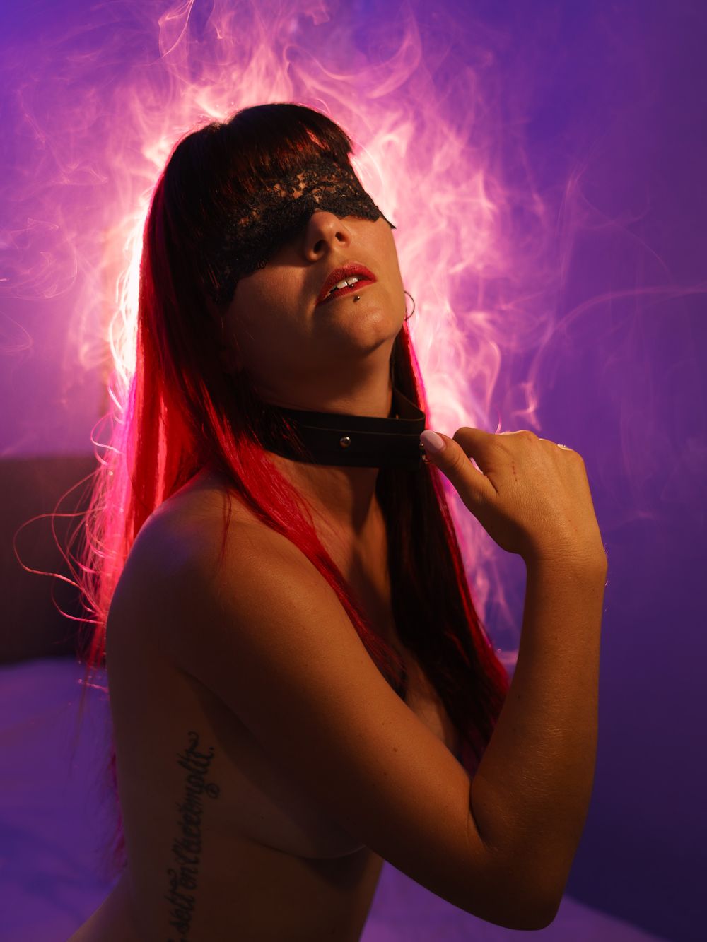 Woman with red hair, lace blindfold, and collar poses dramatically against purple and pink smoky background.