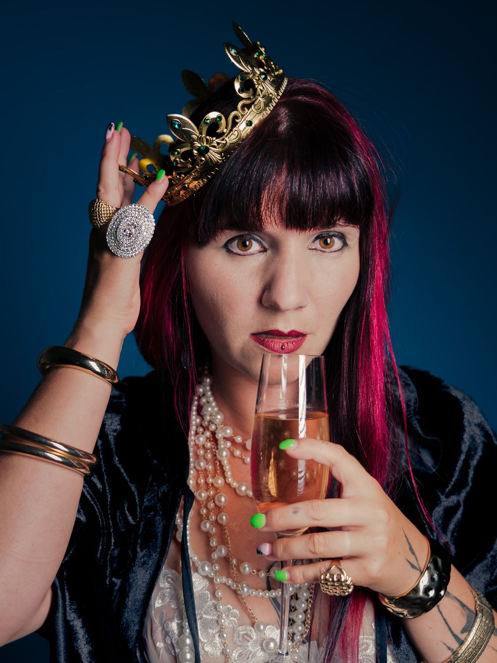Woman with pink hair, gold crown, pearl necklace, and rings sipping champagne, styled in royal-inspired fashion.