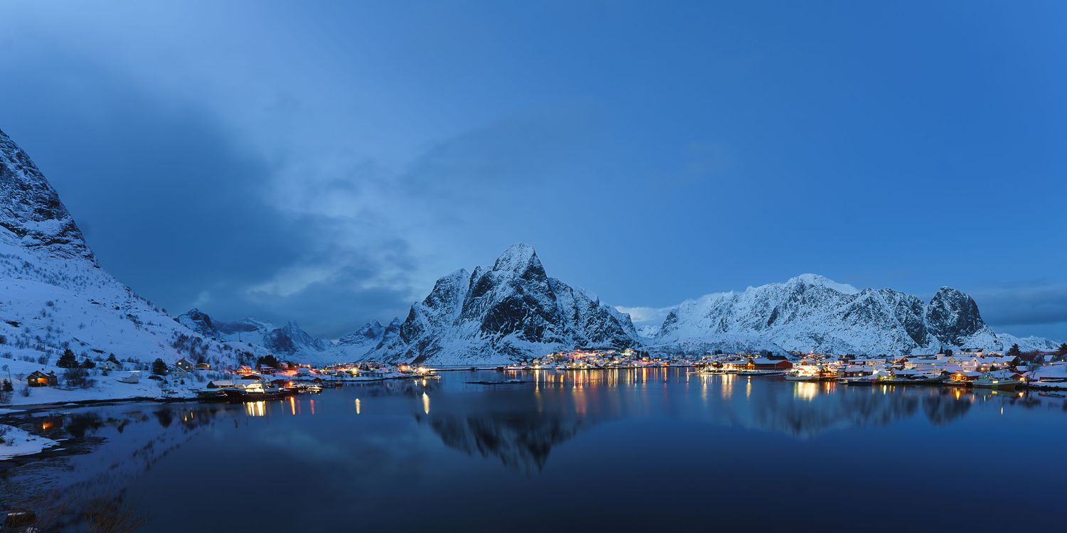 Snow-capped mountains reflect in calm fjord waters at twilight, with a illuminated Norwegian village glowing warmly in Lofoten Islands.