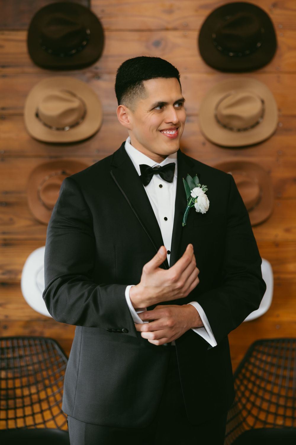 Groom in black tuxedo with white boutonniere smiling, standing in front of wooden wall with hat display.