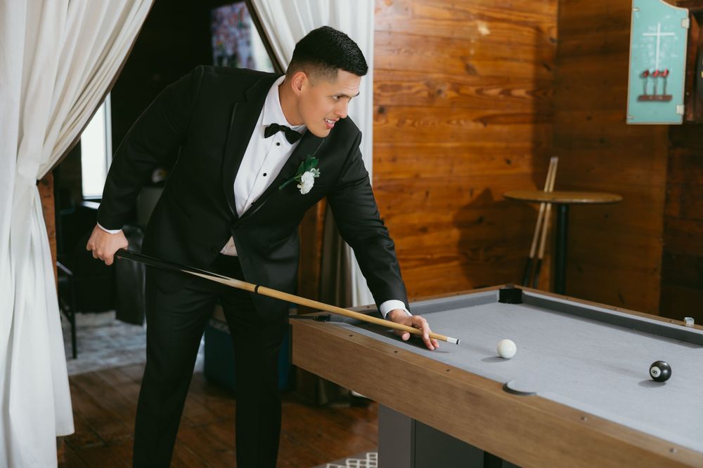 Groom in black tuxedo playing pool at wedding venue, holding cue stick over billiard table.
