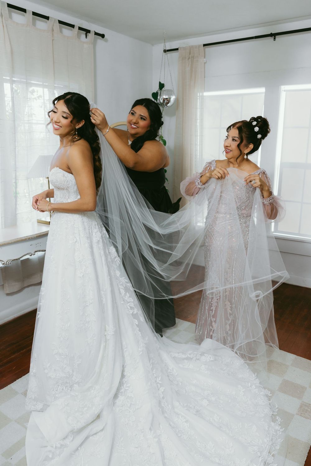 Bride getting veil placed by bridesmaid while another woman holds lace overlay in bright white room.