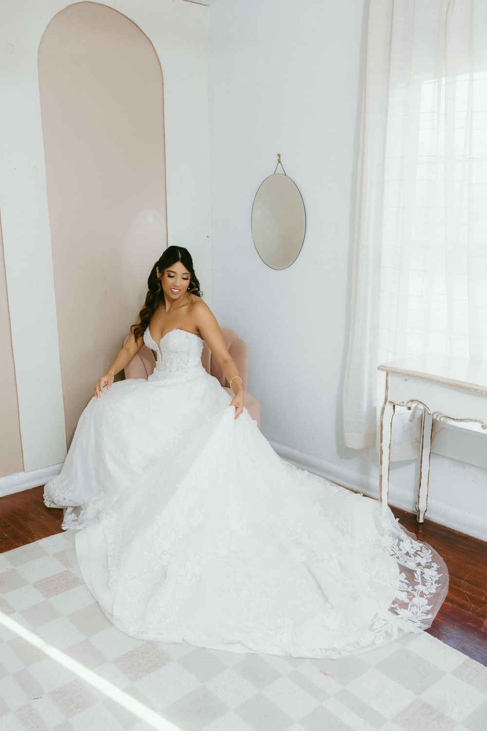 Bride in a strapless ballgown with lace train sits in a bright white bridal suite before her wedding.