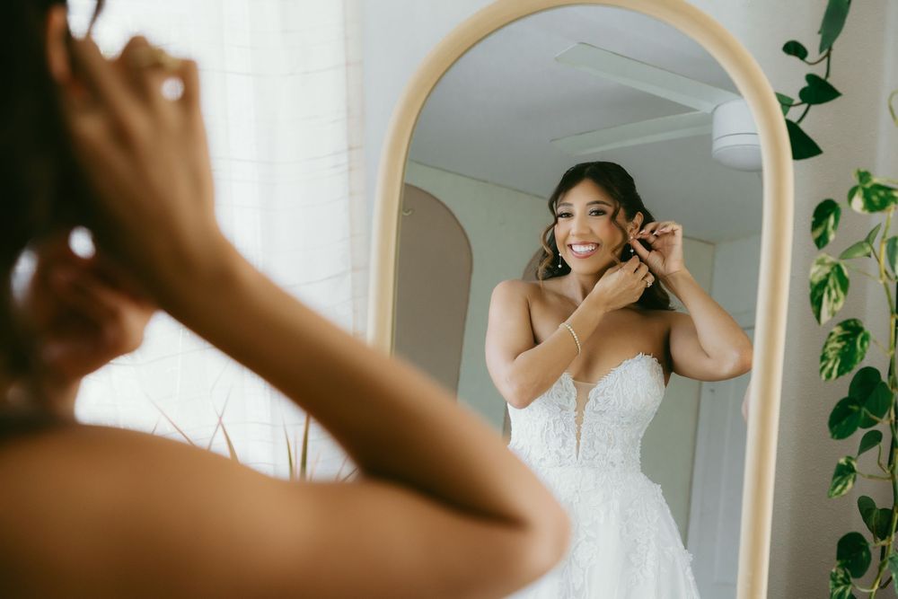 Bride in white lace gown adjusting earring while smiling at her reflection in an arched mirror.
