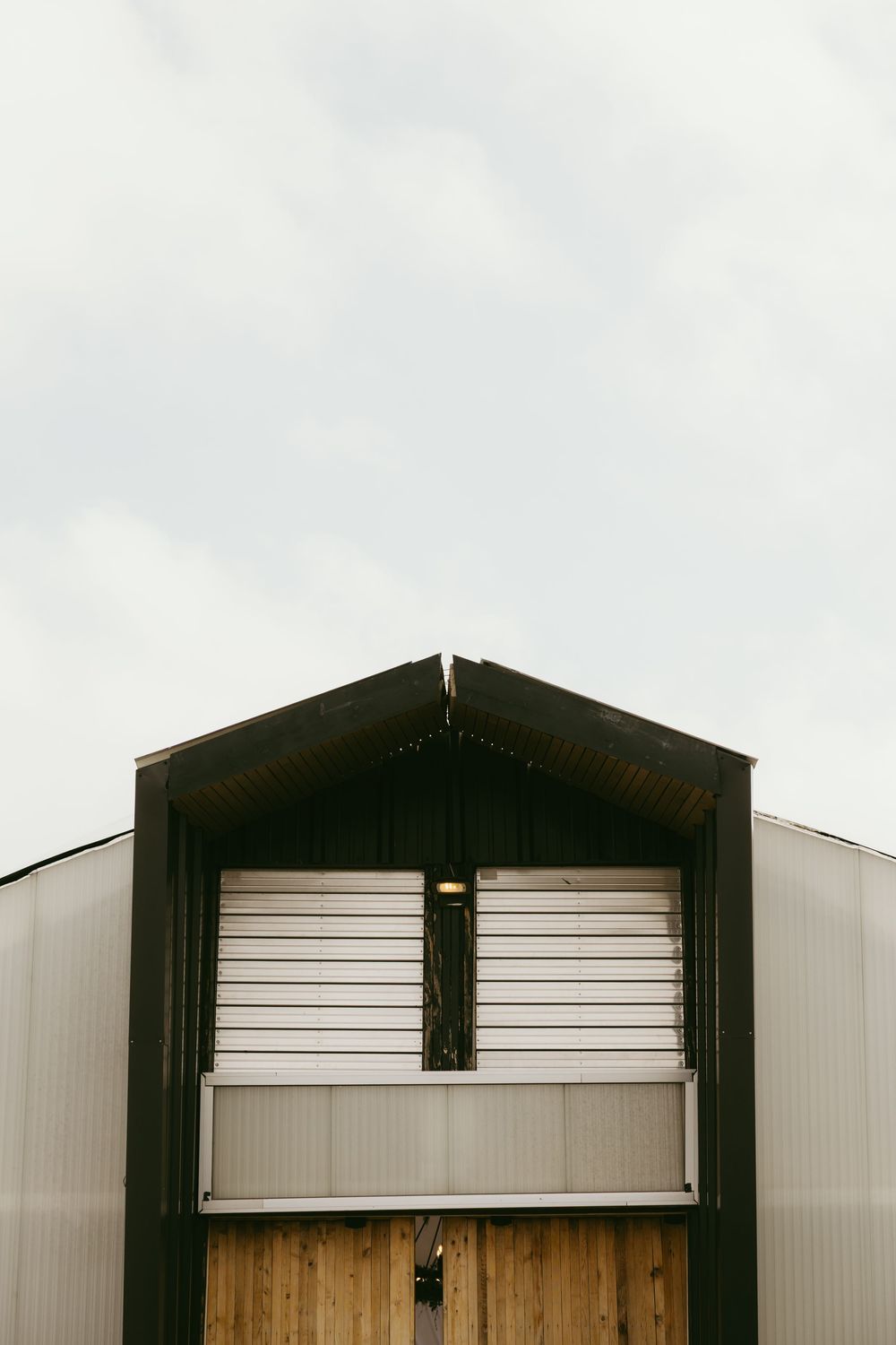 Industrial metal barn building with wooden doors, roller shutter, and dark trim against an overcast sky.