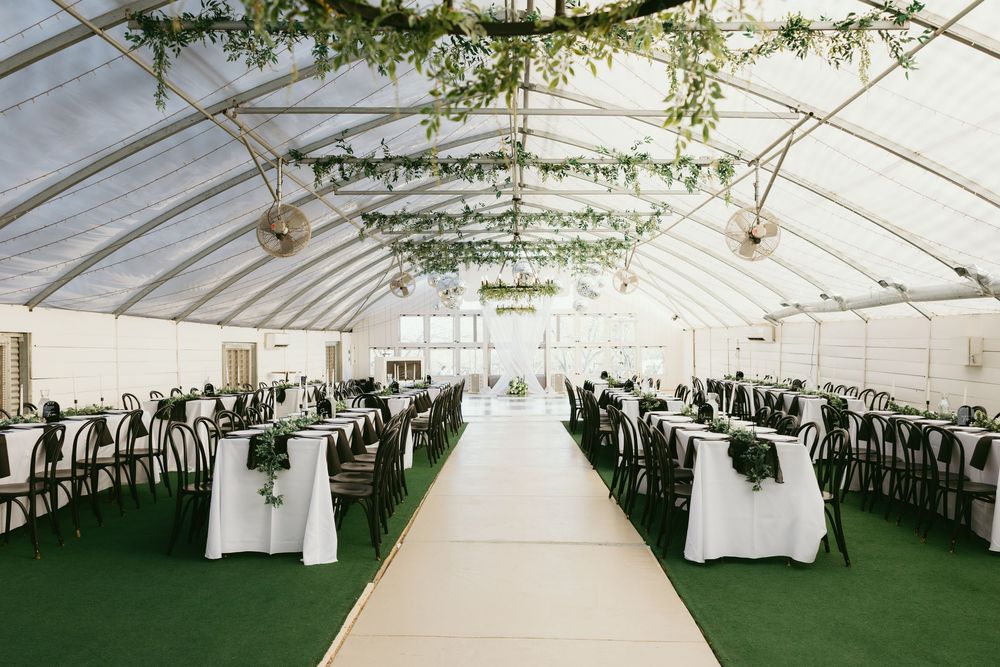 Elegant wedding reception hall under clear tent with long tables, greenery ceiling decor, and white aisle.