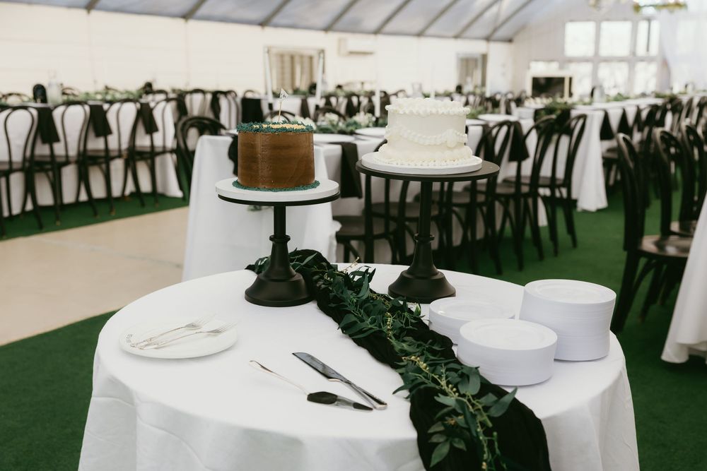 Elegant wedding reception tent with white tablecloths, dark chairs, greenery runners, and tiered cake display.