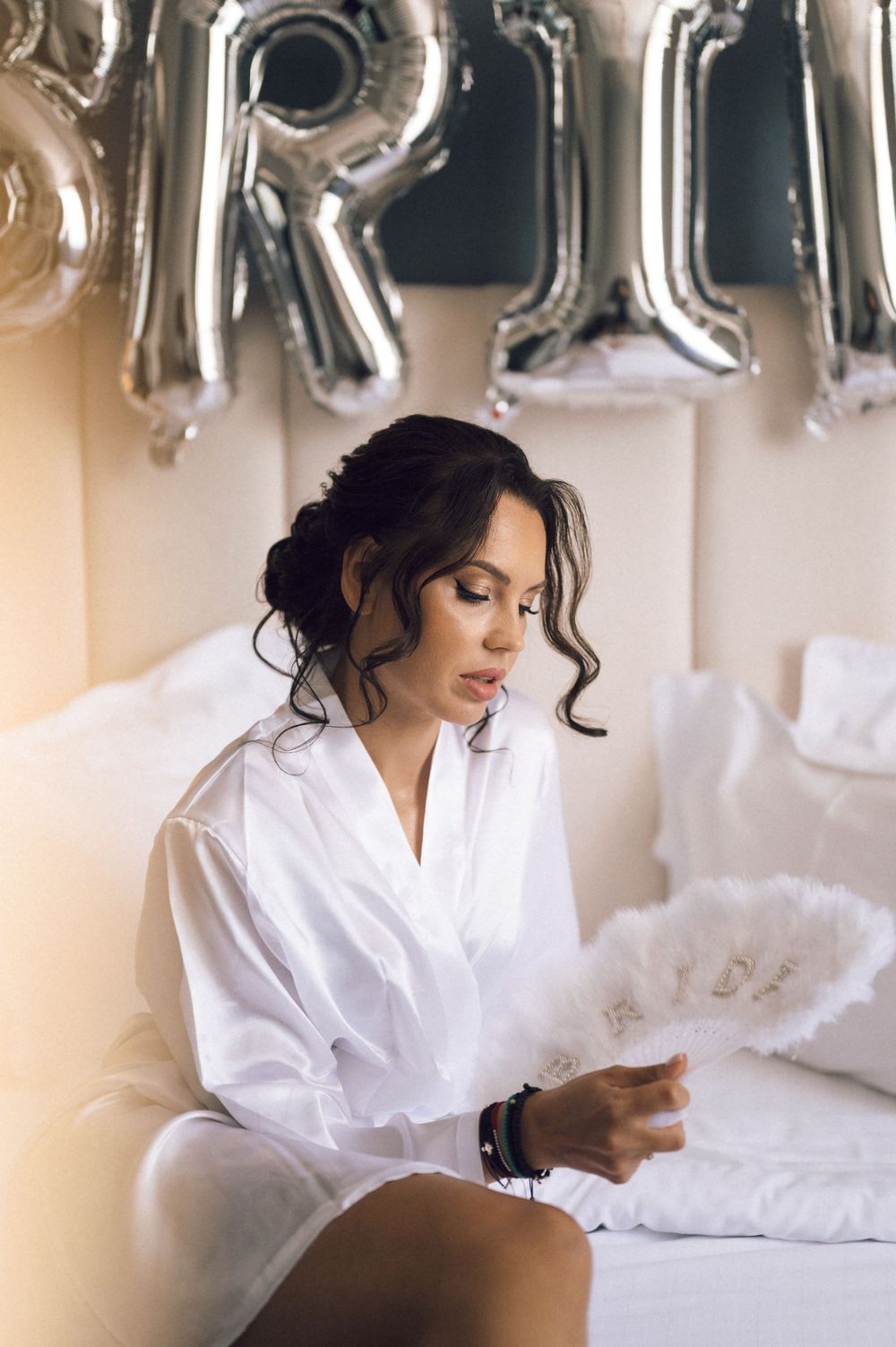 Bride in white robe sitting on bed with silver BRIDE balloons, holding a feather fan during bridal preparations.