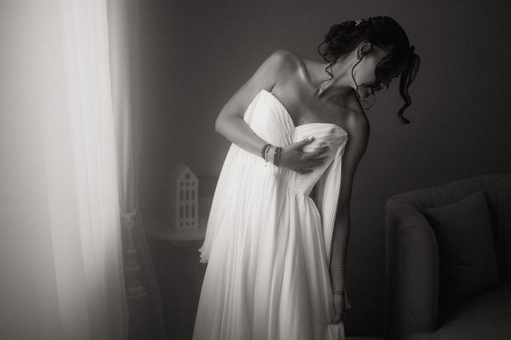 Bride in white strapless wedding gown adjusting dress near window in elegant black and white photo.