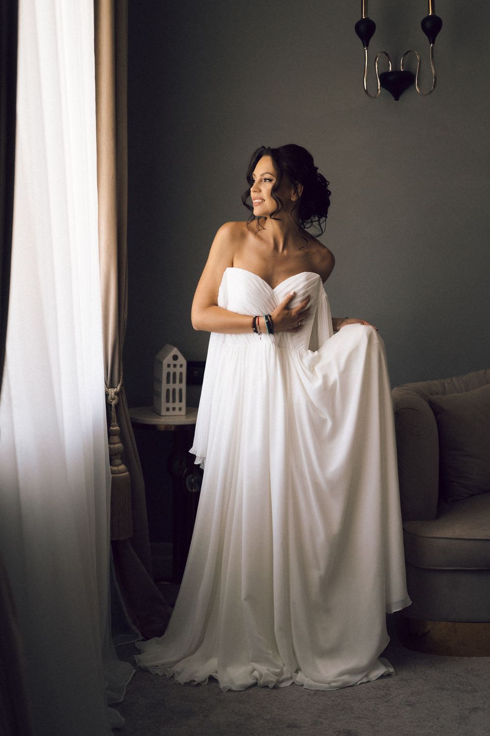 Elegant bride in a strapless white chiffon gown standing by a window in a softly lit room.
