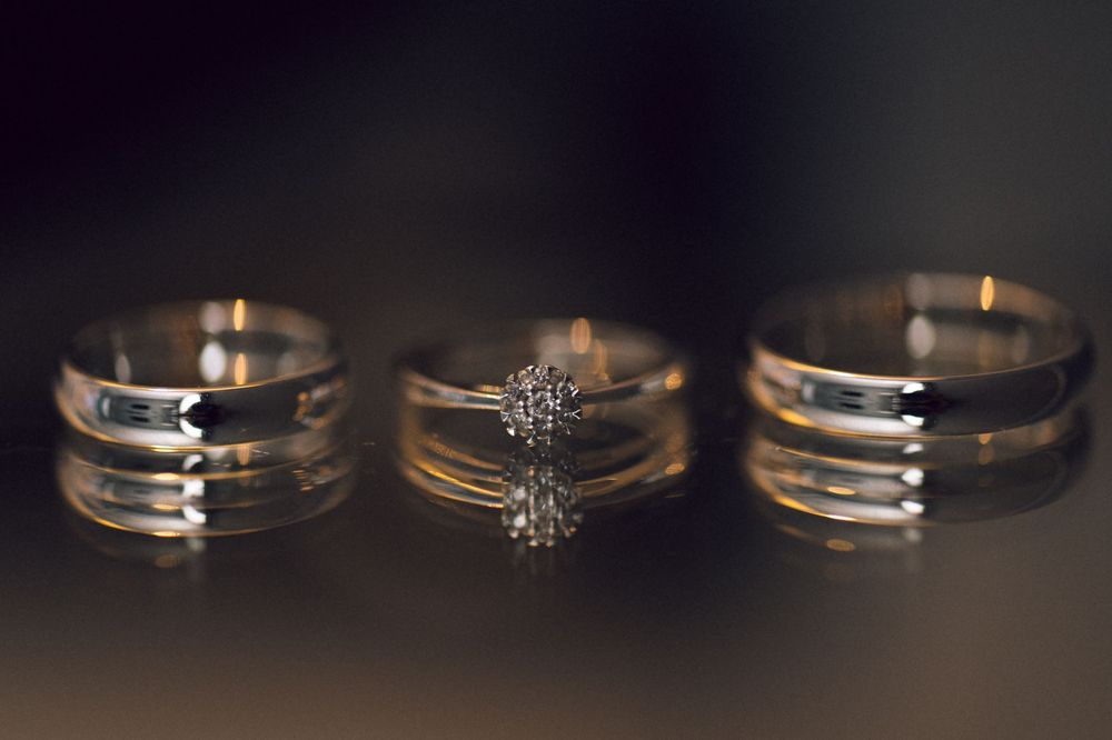 Three gold wedding rings on reflective surface, two plain bands flanking a diamond solitaire engagement ring.