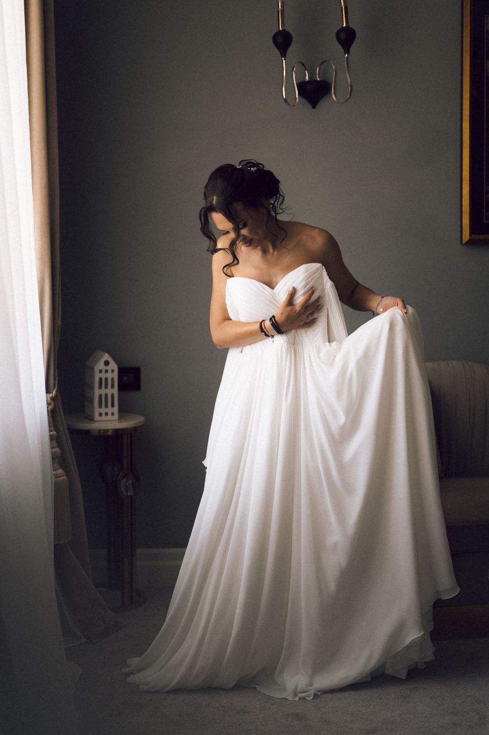 Bride in strapless white wedding gown adjusting her flowing chiffon dress in an elegant room.
