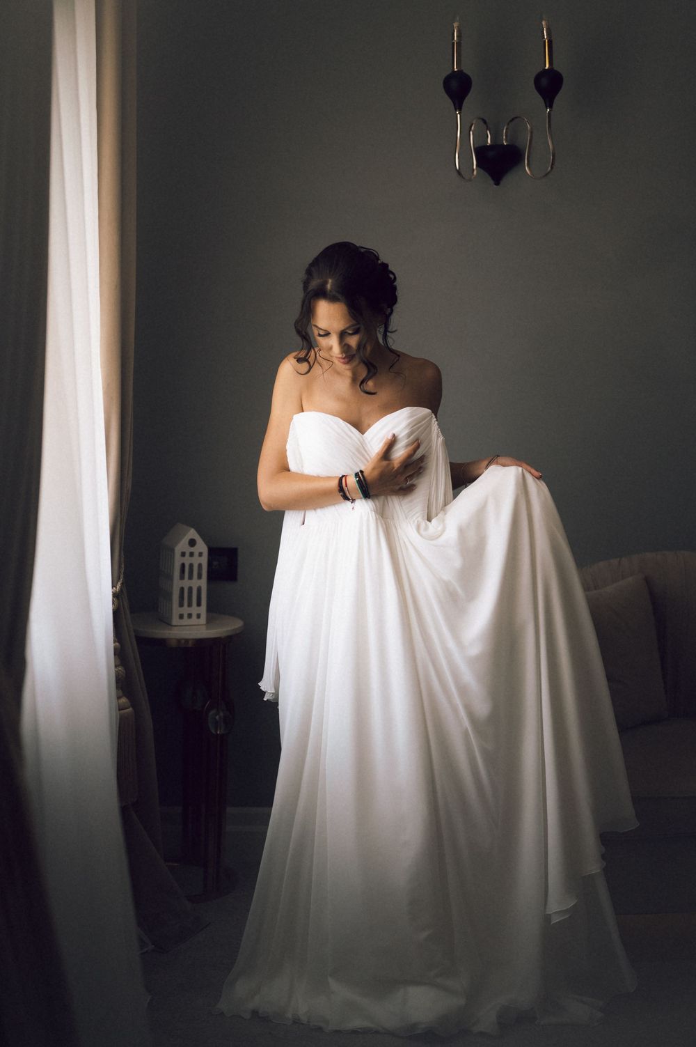 Bride in strapless white chiffon wedding gown adjusting her dress in an elegant dimly lit room.