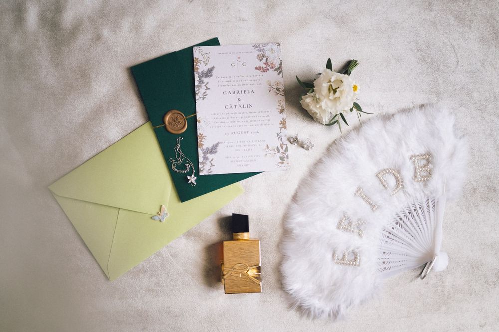 Elegant wedding flat lay with floral invitation, green envelopes, gold perfume bottle, white feather fan, and flower bouquet.