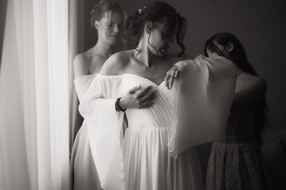 Black and white photo of a bride in a white off-shoulder gown being assisted by bridesmaids near a window.