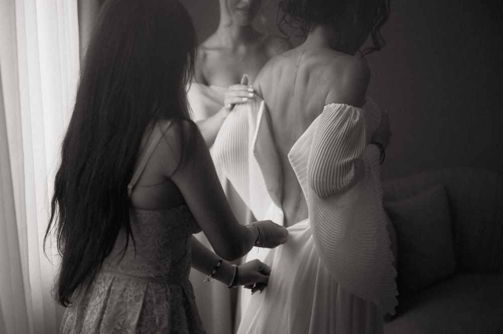 Black and white photo of a bride in a strapless gown being helped by a bridesmaid during wedding preparations.