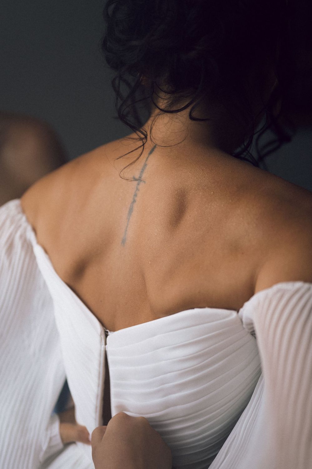 Bride in white off-shoulder gown showing elegant back with delicate cross tattoo on upper spine.