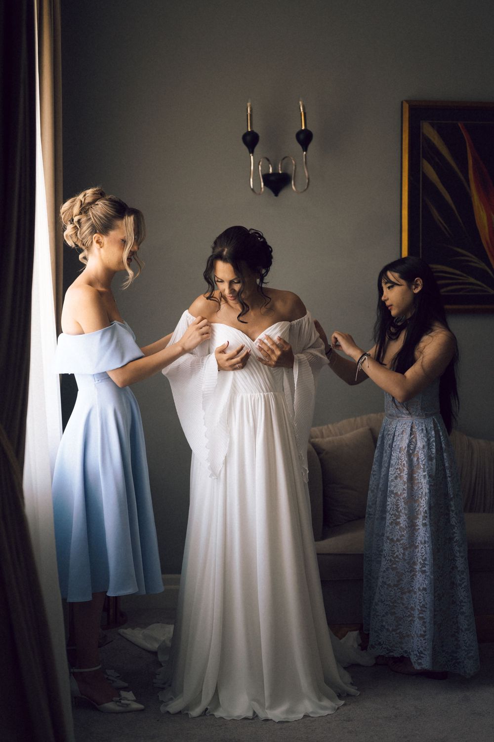 Bride in white off-shoulder gown assisted by two bridesmaids in blue dresses during wedding preparations indoors.