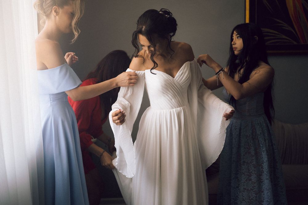 Bridesmaids helping bride into elegant white off-shoulder wedding gown with flowing sleeves in a softly lit room.