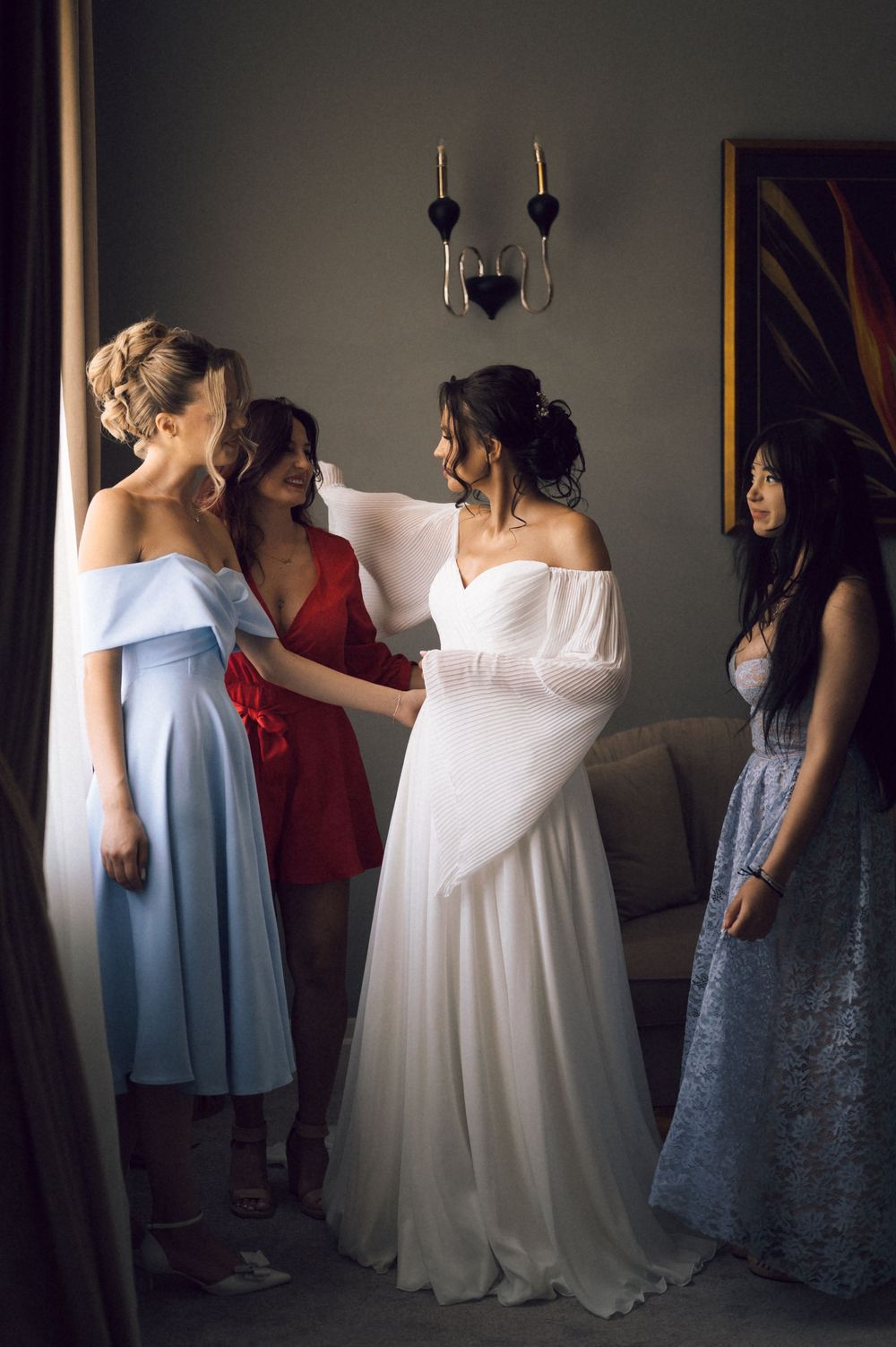 Bride in white off-shoulder gown surrounded by bridesmaids in blue, red, and lace dresses during wedding preparation.