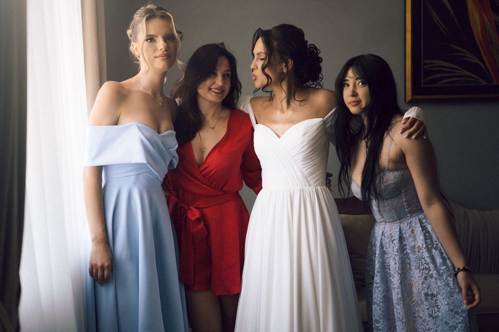 Four women in formal dresses pose together, including a bride in white, for a wedding celebration photo.
