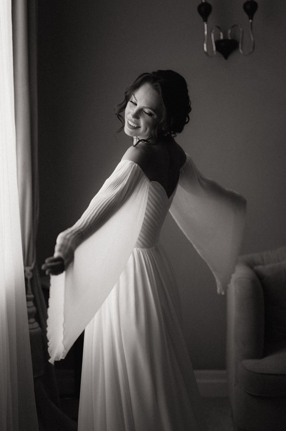 Black and white photo of a smiling bride in an elegant flowing white gown with sheer sleeves, posing gracefully indoors.