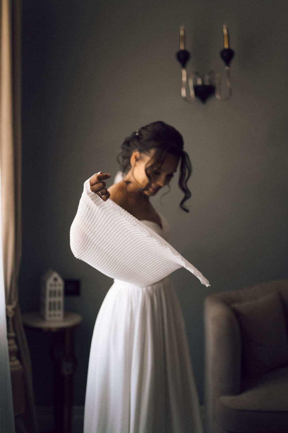 A bride in a white off-shoulder gown holds her dress elegantly in a softly lit room.