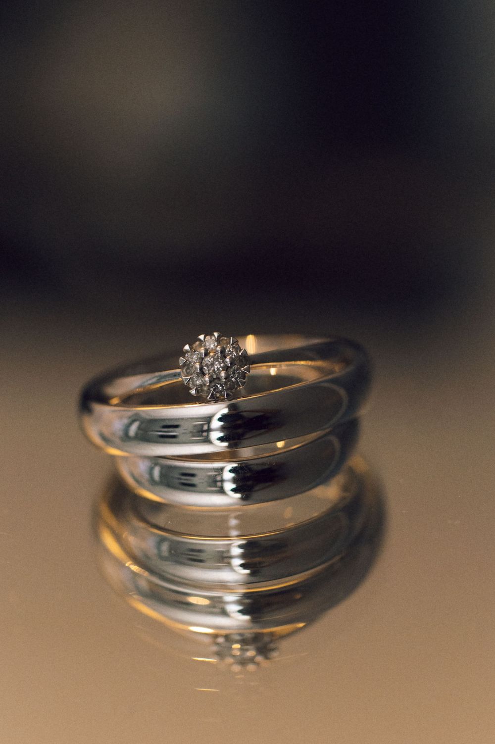 Silver engagement ring with diamond cluster resting on wedding band, reflected on glossy surface against dark background.