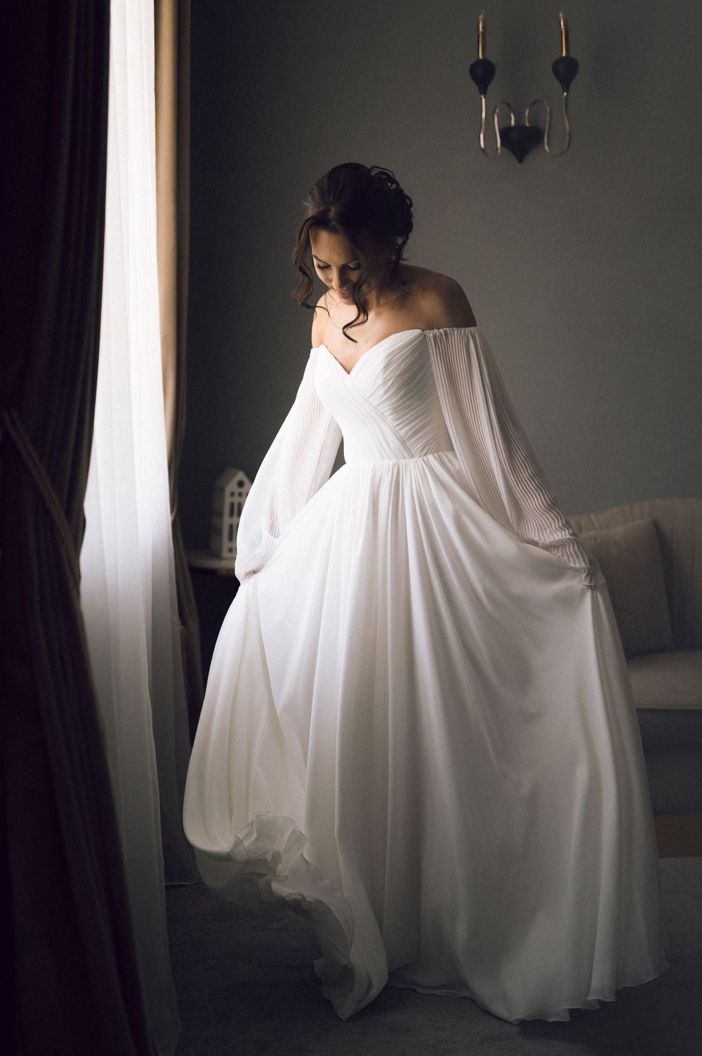 Bride in elegant off-shoulder white chiffon wedding gown with flowing sleeves standing near window in soft light.