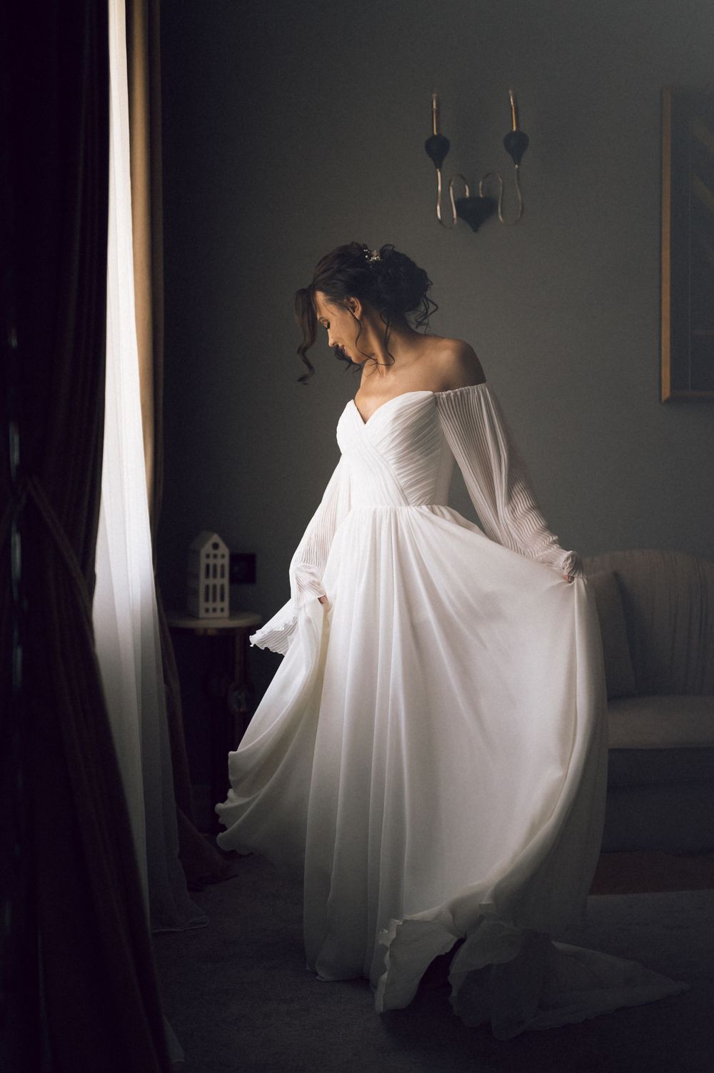Bride in flowing off-shoulder white wedding gown twirling elegantly in a dimly lit room near a window.