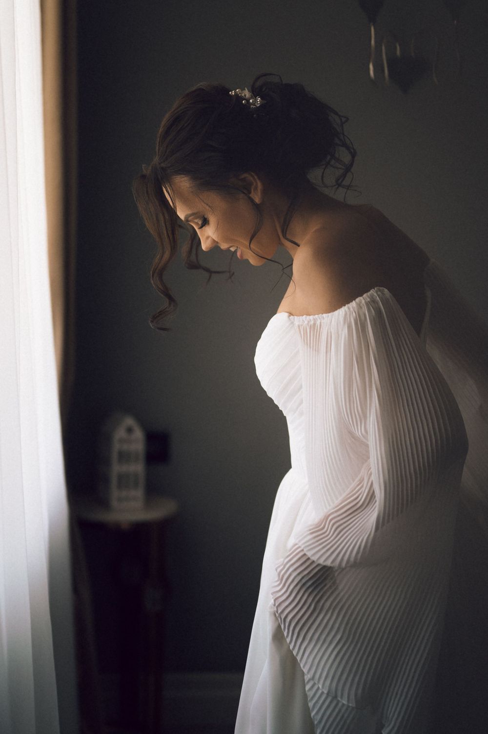Bride in white pleated strapless gown standing by window light, head bowed, hair adorned with floral pins.