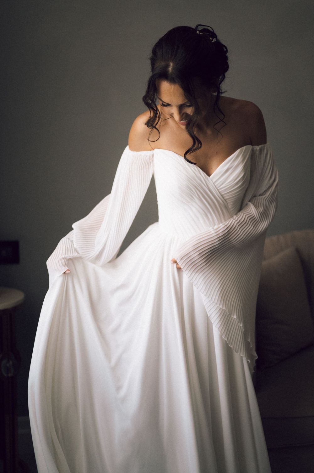 Bride in elegant off-shoulder white wedding gown with long bell sleeves, looking down gracefully in soft studio lighting.
