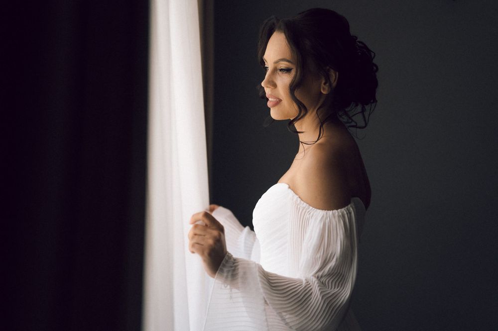 Elegant woman in white off-shoulder top gazing out a window with sheer curtains in soft natural light.