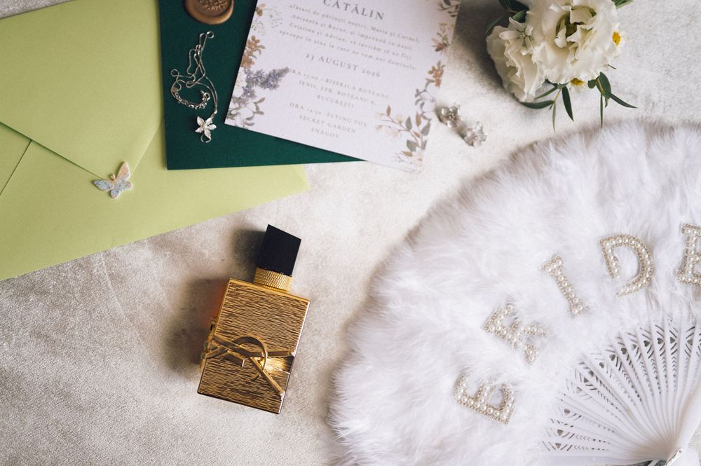 Elegant wedding flat lay with gold perfume bottle, green envelope, floral invitation, pearl fan, and delicate jewelry.