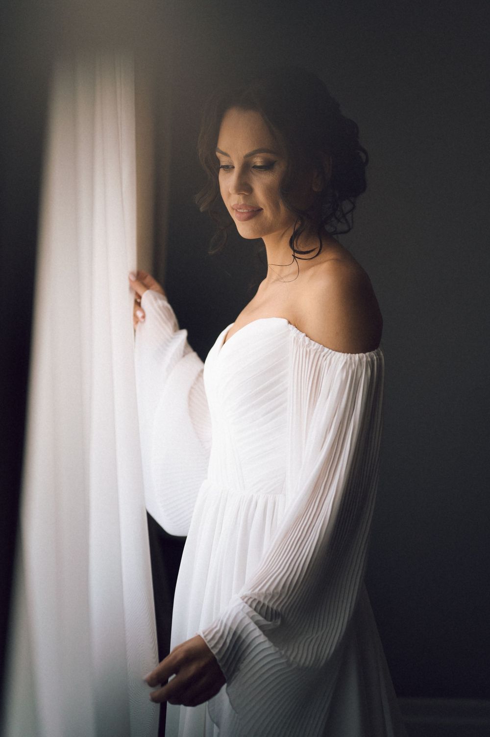 Elegant bride in white off-shoulder gown with flowing sleeves stands by window in soft natural light.