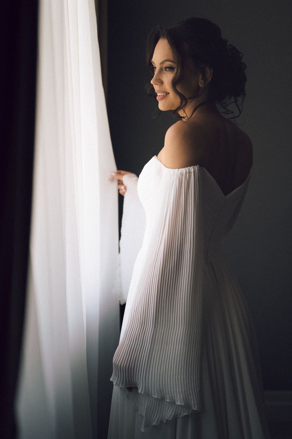 Elegant bride in white off-shoulder pleated gown smiling by sheer curtains in soft natural light.