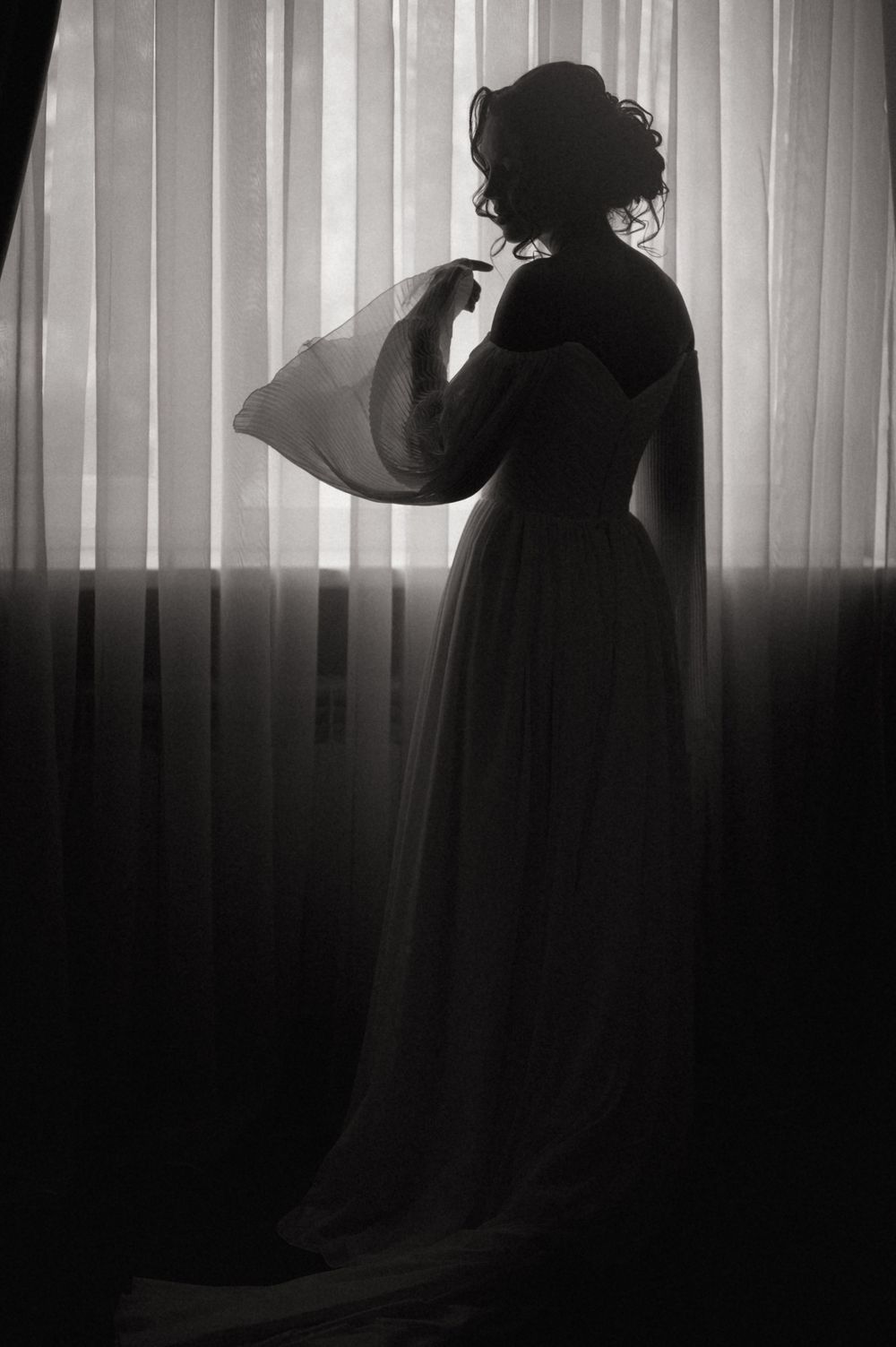 Black and white silhouette of a woman in an elegant gown standing by sheer curtains in soft backlight.