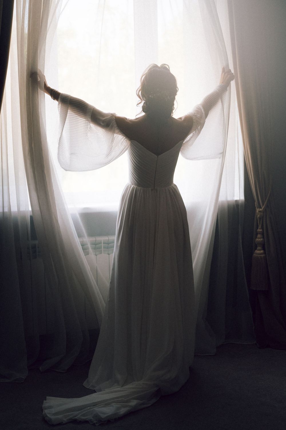 Bride in white wedding gown standing at sunlit window, pulling sheer curtains apart in elegant backlit silhouette.