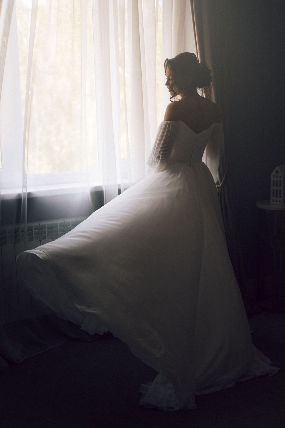Elegant bride in a flowing white wedding gown standing by a sunlit window in a softly lit room.