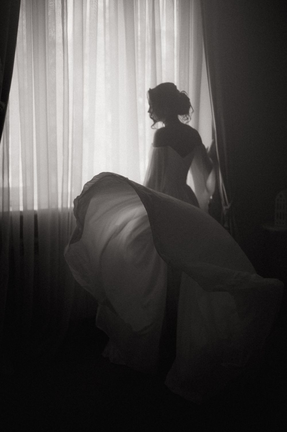 Black and white silhouette of a bride in flowing wedding gown standing by sheer curtained window in dramatic light.