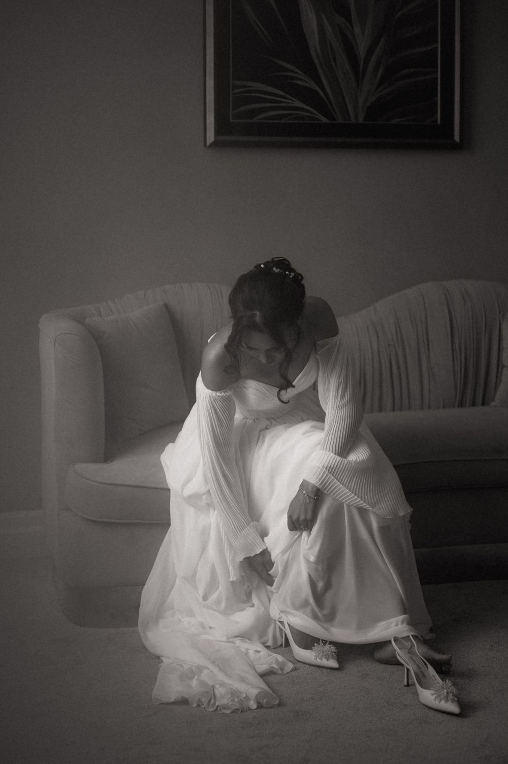 Black and white photo of a bride in a white gown sitting on a sofa, head in hands, wearing lace gloves.