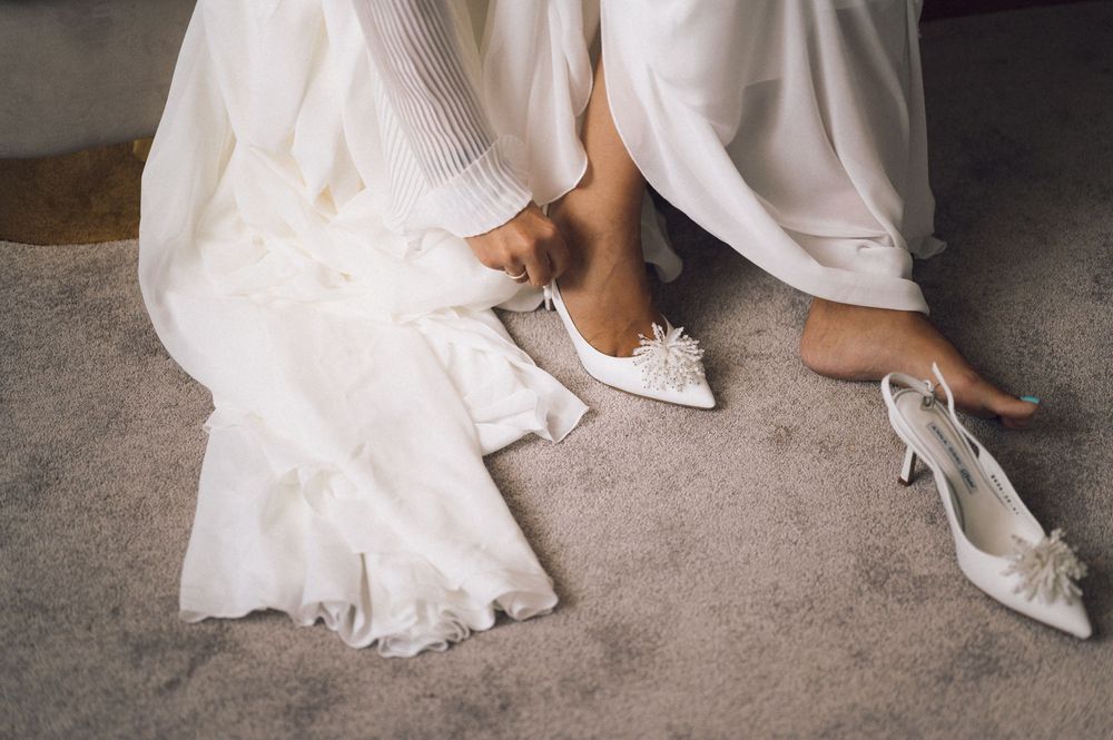 Bride putting on white lace heels while sitting in wedding dress on carpeted floor.