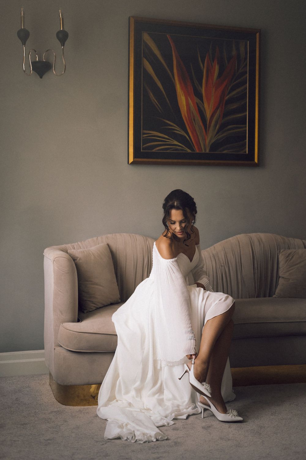 Bride in white wedding gown adjusting her heel on a beige sofa, with elegant wall art and sconce in background.