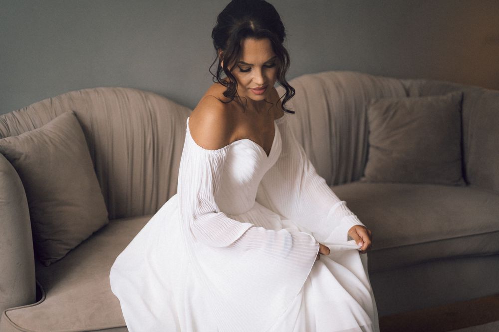 Bride in off-shoulder white gown sitting gracefully on a beige sofa, looking down elegantly.
