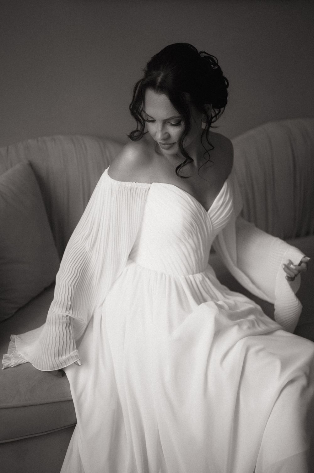 Black and white photo of a bride in an elegant off-shoulder white gown sitting gracefully on a sofa.