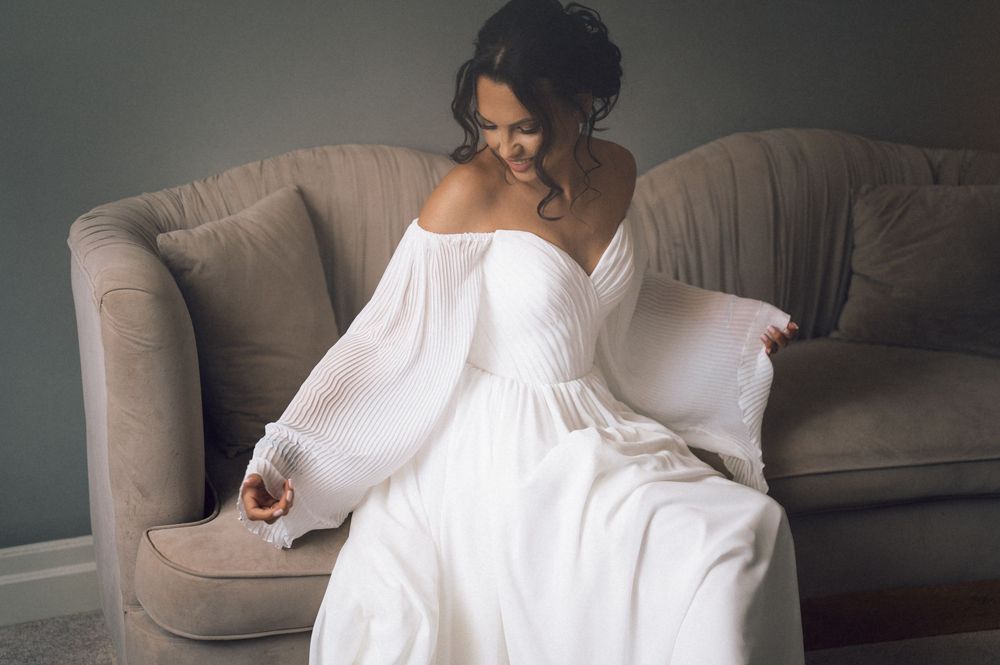 Bride in white off-shoulder gown sitting on beige sofa, smiling down at her dress.