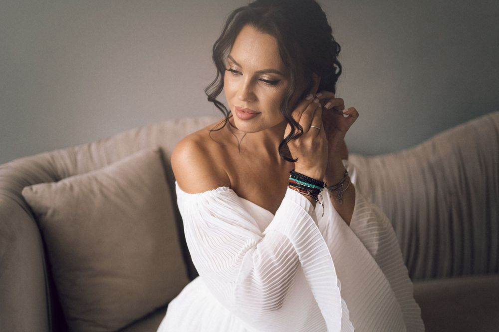 Elegant woman in white off-shoulder sweater sitting on a beige sofa, wearing a black smartwatch, looking down pensively.