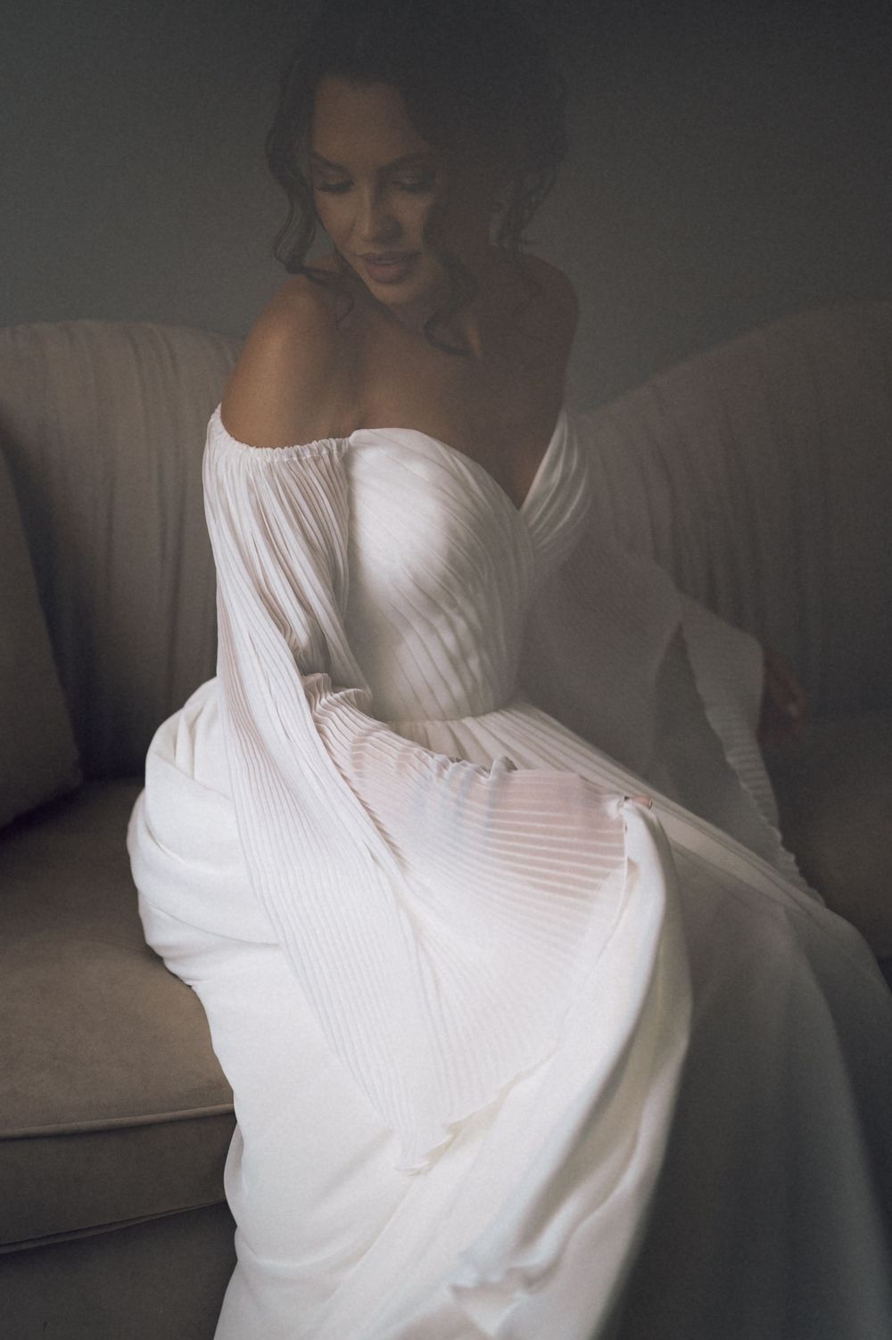 Bride in elegant white off-shoulder pleated wedding gown seated gracefully on a neutral sofa in soft moody lighting.