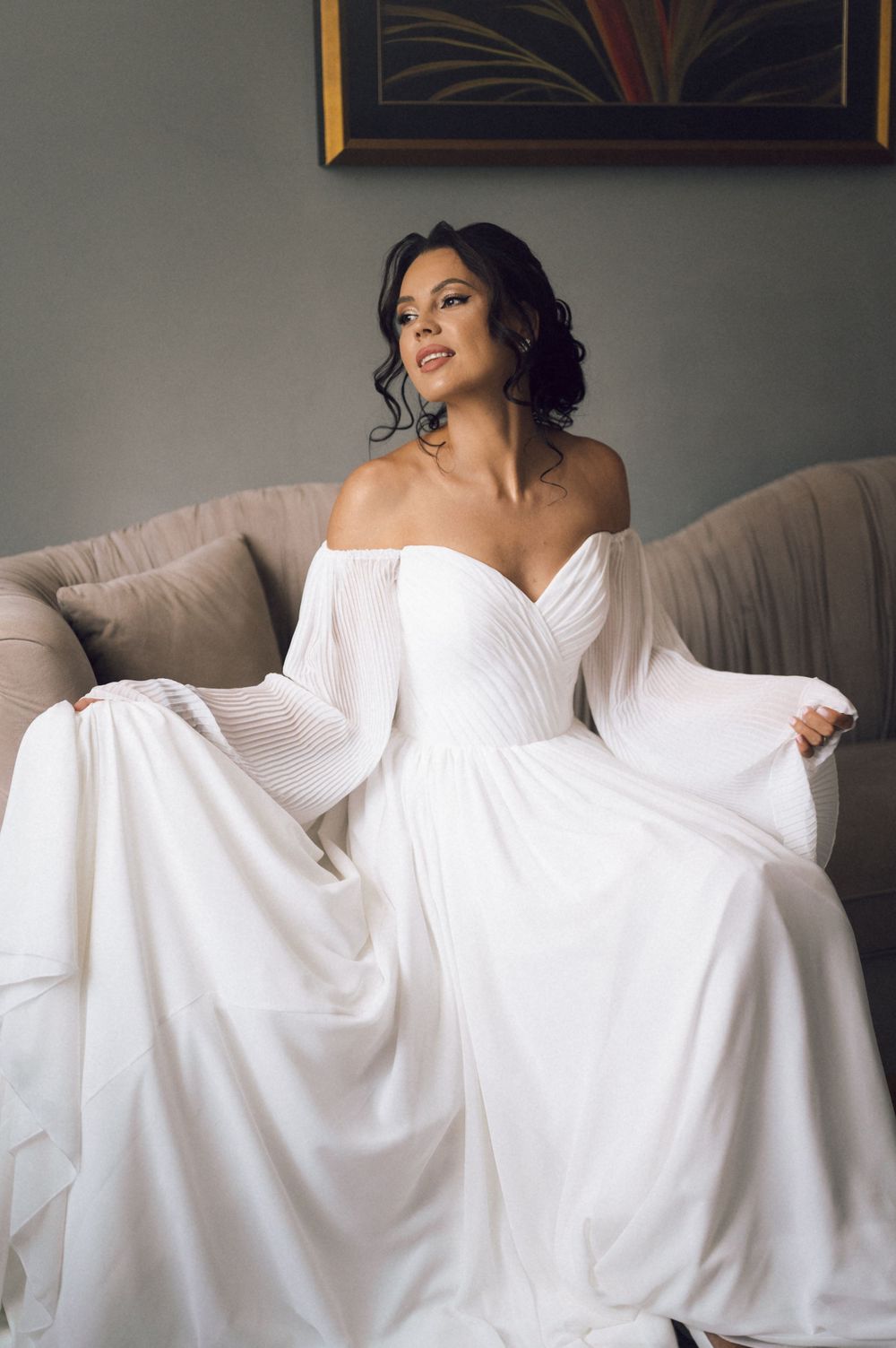 Bride in an off-shoulder white wedding gown with flowing sleeves sits gracefully on a beige sofa, smiling softly.